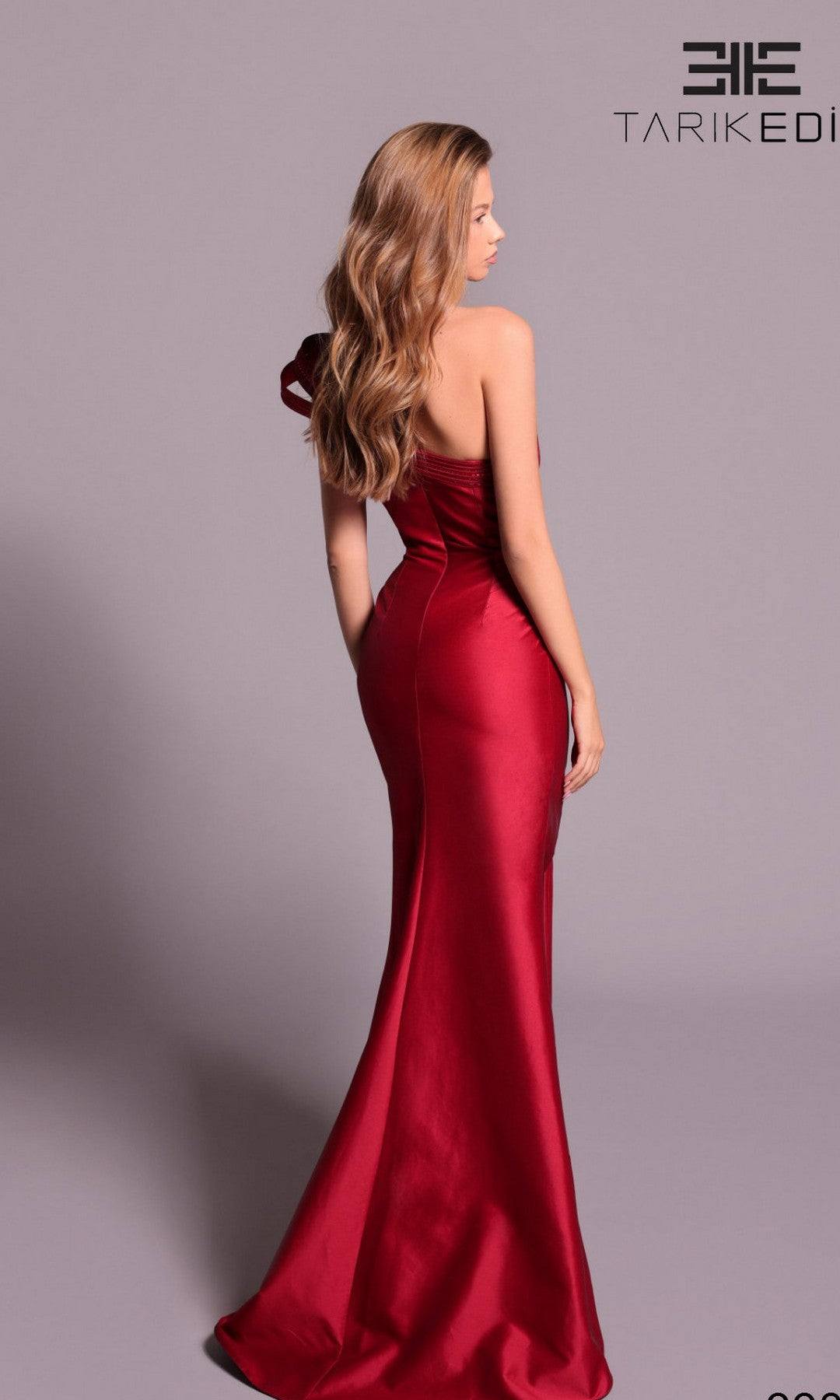 A woman with long wavy hair models the Tarik Ediz 99065 long formal dress— a fitted, floor-length gown with a one-shoulder strap and high leg slit— against a plain light background.